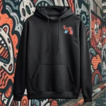 Front view of black pullover hoodie featuring a cow juggling spices and ingredients for an exotic soup with a bandaid on its crotch.