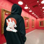 A male model with his back turned wearing a black Manifesto hoodie featuring a giant bald man crawling with its hands and feet as multiple eyes spew from its chest and chin