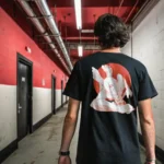 A male model with his back turned wearing a black Manifesto shirt featuring a giant bald man crawling with its hands and feet as multiple eyes spew from its chest and chin