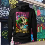 Back view of black Tropic Soul hoodie displaying a man relaxing on a hammock tied to the Skull Island's palm trees next to a giant wave rear graphic placement and hood detail.