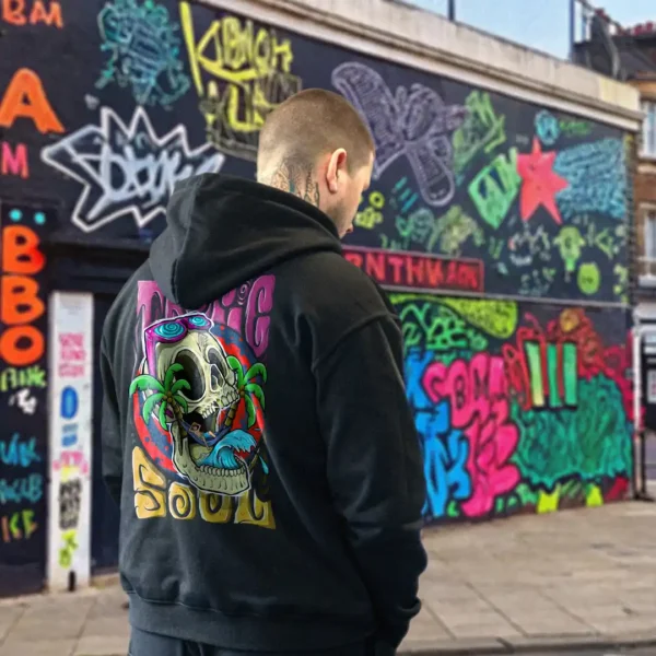 A male model with his back turned wearing a black tropic soul hoodie featuring a man relaxing on a hammock tied to the Skull Island's palm trees next to a giant wave graphic.