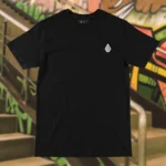 Front view of black tshirt featuring a man relaxing on a hammock tied to the Skull Island's palm trees next to a giant wave graphic.