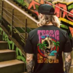 A male model with his back turned wearing a black tropic soul tshirt featuring a man relaxing on a hammock tied to the Skull Island's palm trees next to a giant wave graphic.