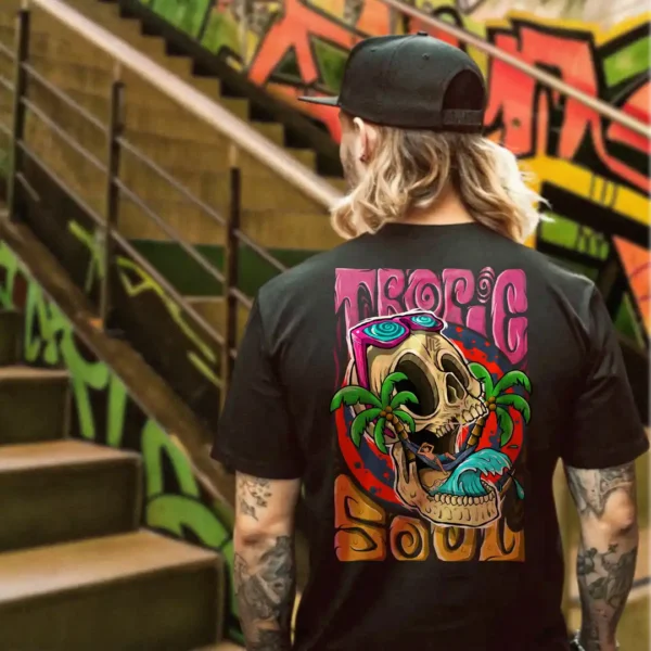 A male model with his back turned wearing a black tropic soul tshirt featuring a man relaxing on a hammock tied to the Skull Island's palm trees next to a giant wave graphic.