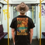 A male model with his back turned wearing a black tropic soul tshirt featuring the Sandset beach and waves graphic.