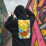 A male model with his back turned wearing a black Easy Days hoodie featuring a roasted pig skewered by a bamboo stick, lying underneath an umbrella taking cover from the sun.