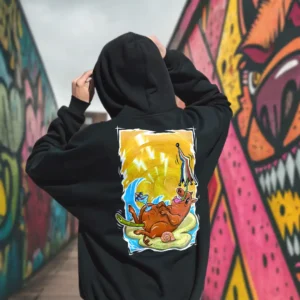 A male model with his back turned wearing a black Easy Days hoodie featuring a roasted pig skewered by a bamboo stick, lying underneath an umbrella taking cover from the sun.