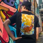 A male model with his back turned wearing a black Easy Days tshirt featuring a roasted pig skewered by a bamboo stick, lying underneath an umbrella taking cover from the sun.