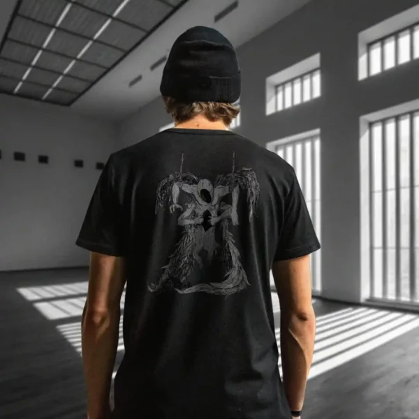 A male model with his back turned wearing a black Manifesto shirt featuring a gray winged creature of dark, visibly hanged by ropes, opening its stomach with its unseen face